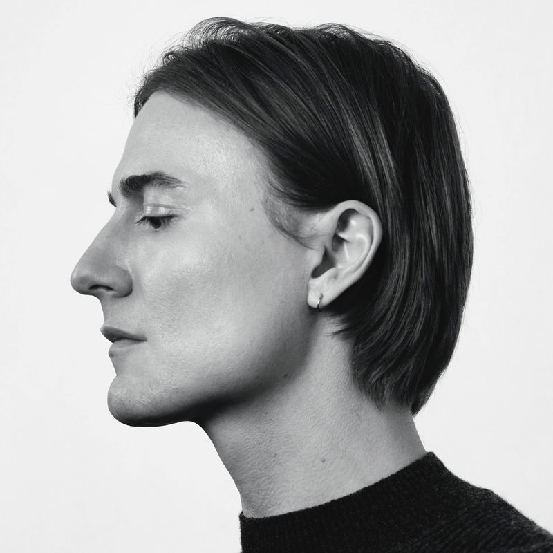 Black and white profile portrait of Alain Paul in a dark high-neck knit sweater against a plain white background.