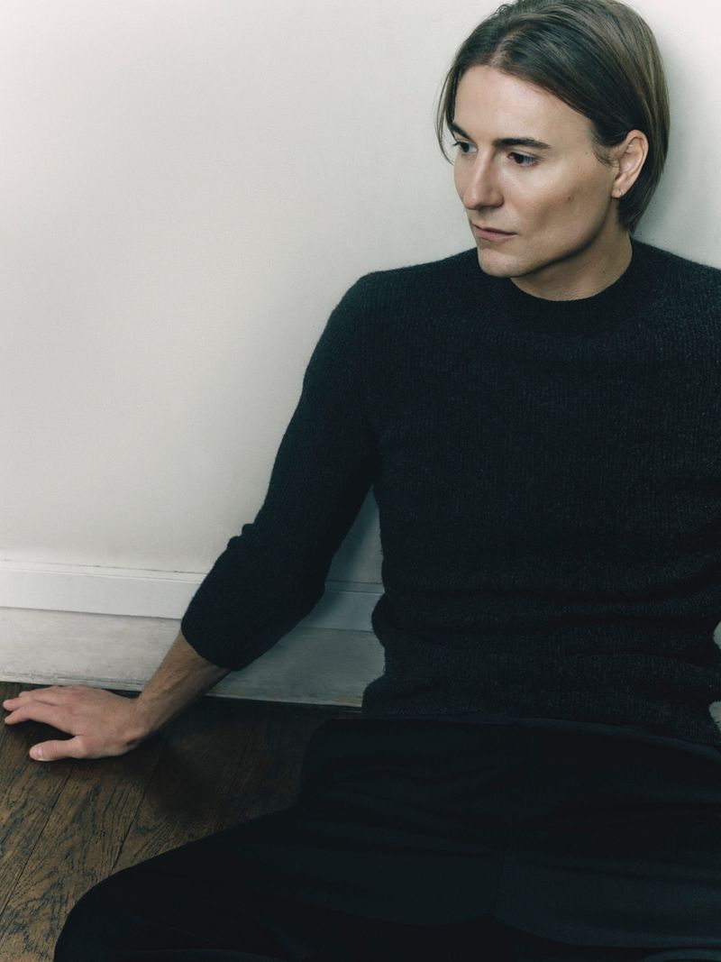 Alain Paul wearing a dark long-sleeve knit sweater, seated on a dark wooden floor against a plain wall.