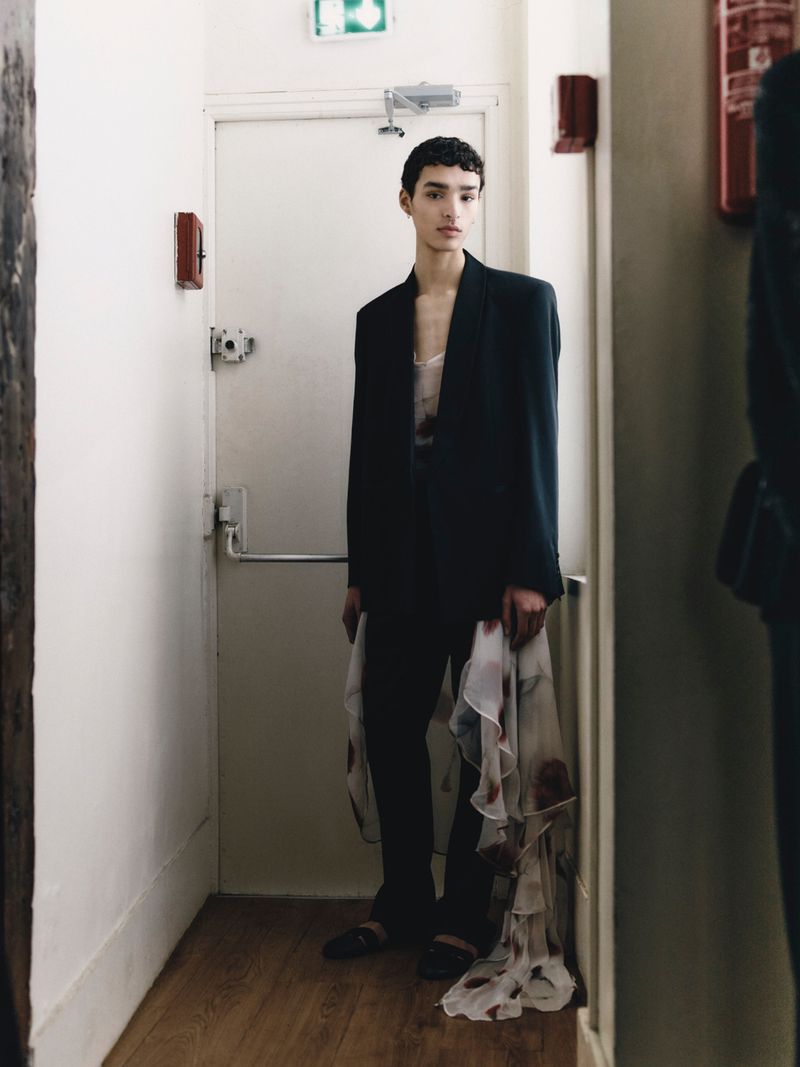 Model wearing a tailored black blazer and trousers with draped sheer printed fabric standing inside a doorway.