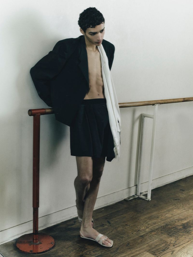A male model in an open black blazer and shorts standing against a ballet barre in a dance studio.