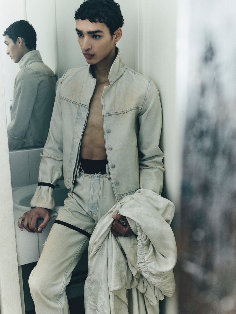Man in a washed off-white denim jacket and matching trousers posing near a bathroom mirror while holding draped fabric.