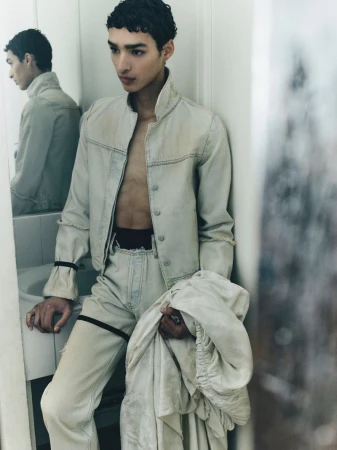 Man in a washed off-white denim jacket and matching trousers posing near a bathroom mirror while holding draped fabric.