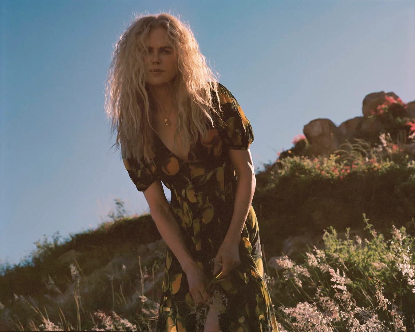 Adult female with blonde curly hair wearing a floral dress, standing outdoors in a field on a sunny day with a mountain in the distance.
