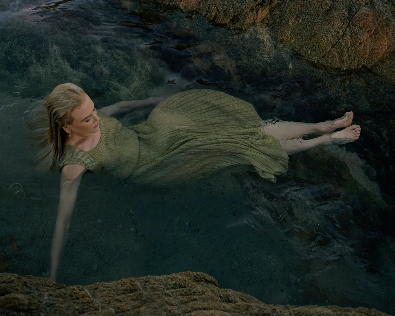 Woman in a green dress lies semi-submerged in dark, dappled ocean water surrounded by smooth, tan rocks in an outdoor setting.
