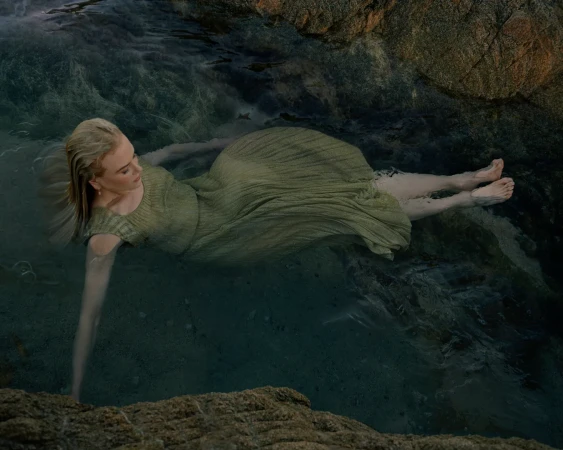 Woman in a green dress lies semi-submerged in dark, dappled ocean water surrounded by smooth, tan rocks in an outdoor setting.