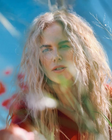 Close-up portrait of a woman with long blonde curly hair in an outdoor setting with a blurred background of blue and red colors.