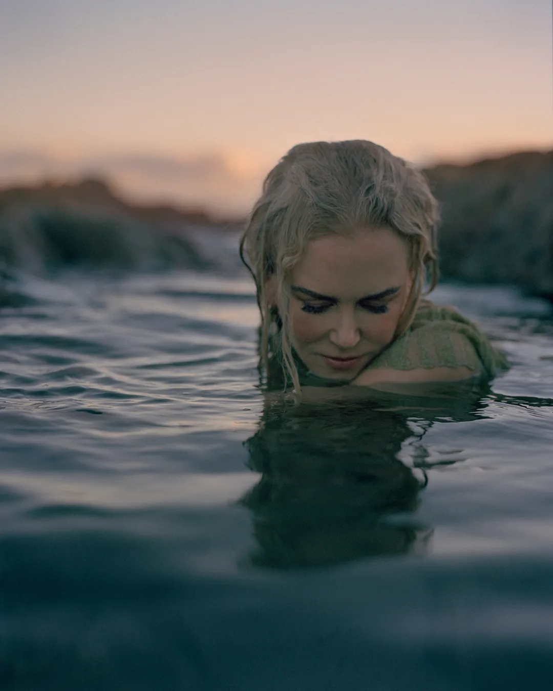 Woman with light skin and blonde wavy hair in water, looking down, with blurred rock formations in the background during sunset.