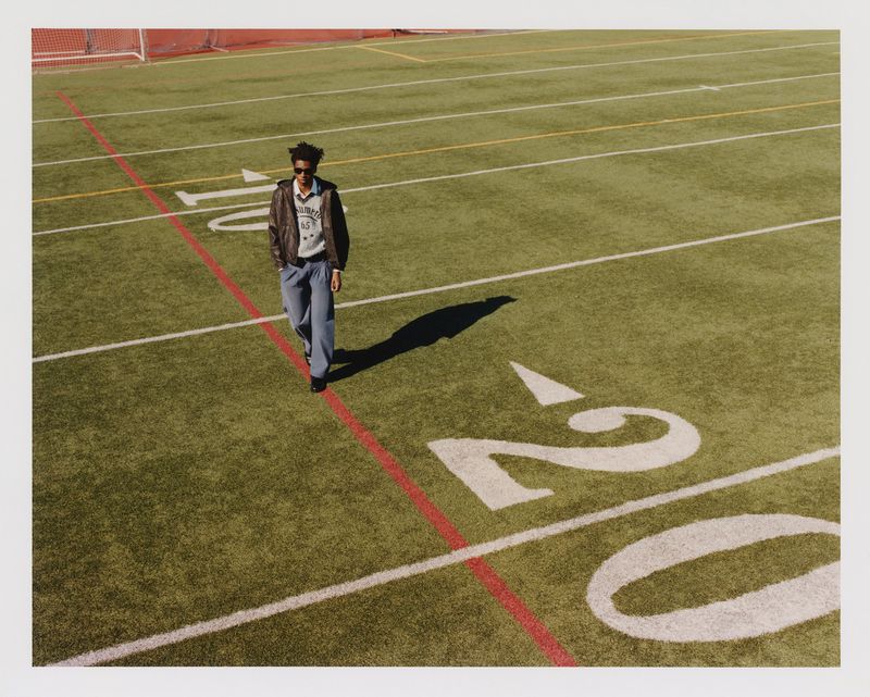 Adult male in casual wear walks on an outdoor football field on a sunny day with bright natural lighting, wearing jeans and a bomber jacket.