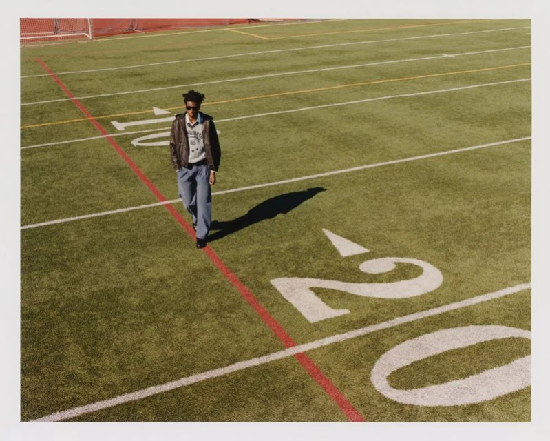 Adult male in casual wear walks on an outdoor football field on a sunny day with bright natural lighting, wearing jeans and a bomber jacket.
