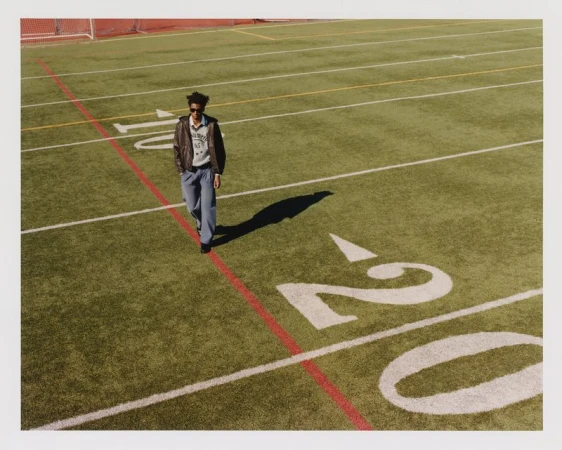 Adult male in casual wear walks on an outdoor football field on a sunny day with bright natural lighting, wearing jeans and a bomber jacket.