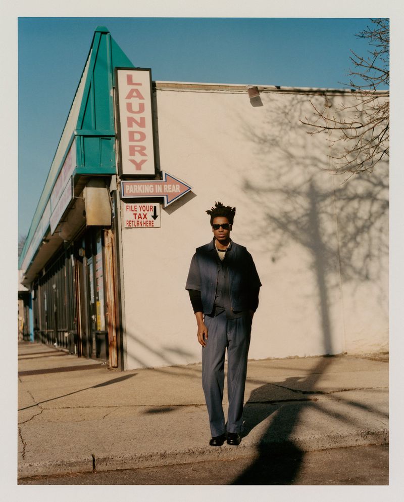 Adult male in a blue suit and sunglasses standing on a city sidewalk with a laundromat in the background on a sunny day.