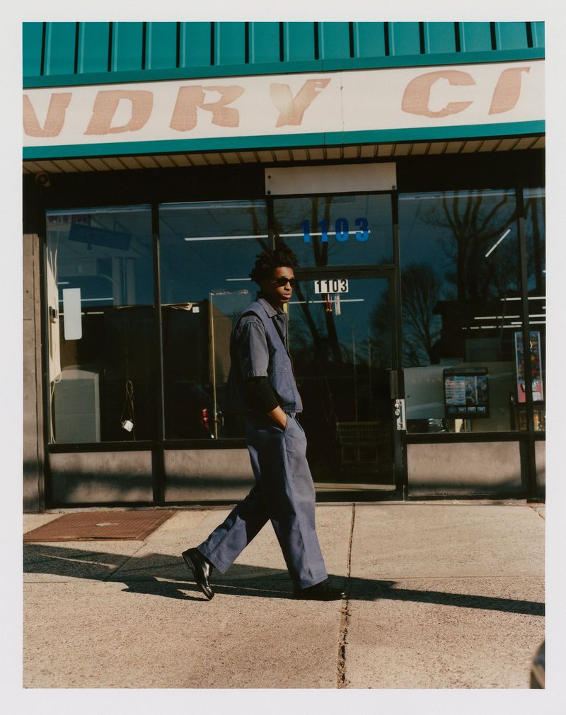 A man in a blue jumpsuit and sunglasses walks past a laundry business on a sunny day, reflected in the storefront's glass windows.