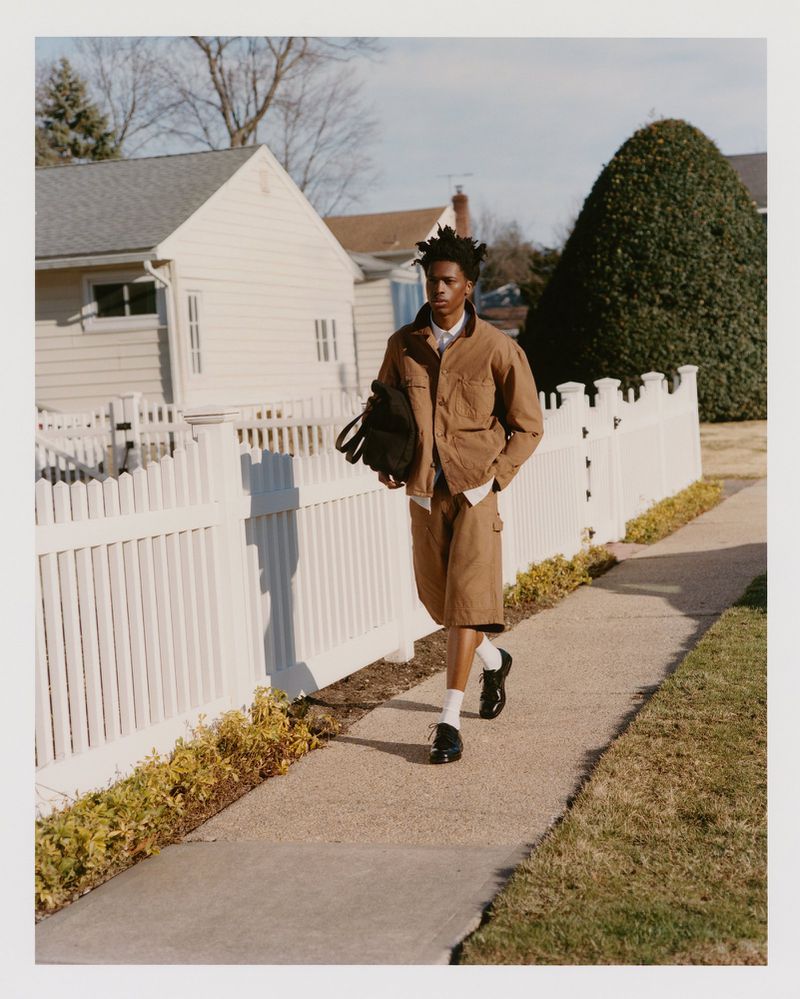 An African American man walks along a sidewalk in front of a white picket fence, wearing a brown jacket, shorts, and white socks.