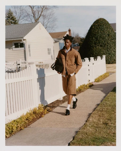 An African American man walks along a sidewalk in front of a white picket fence, wearing a brown jacket, shorts, and white socks.