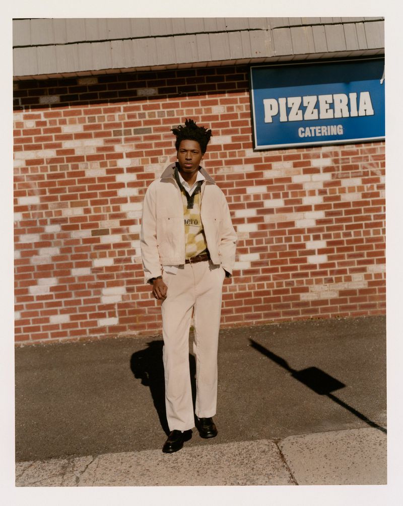 Adult male in white jacket and pants stands on sidewalk in front of brick building with pizzeria sign; shadow visible on ground.