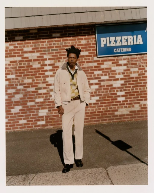 Adult male in white jacket and pants stands on sidewalk in front of brick building with pizzeria sign; shadow visible on ground.