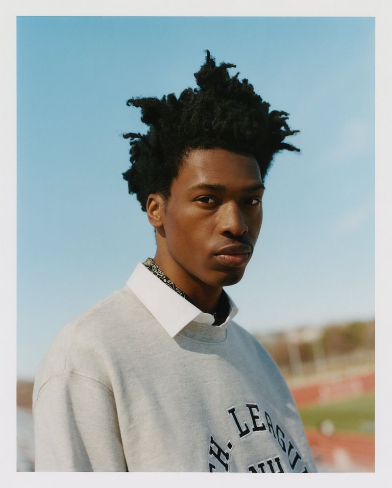 Portrait of a young adult African-American man in a grey sweatshirt and white collar, against a bright blue sky and blurry background.