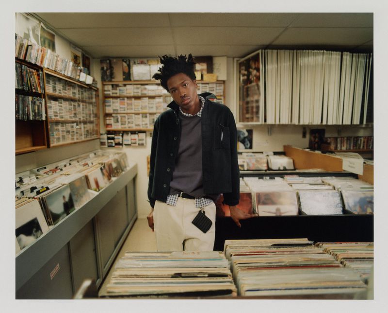 Young adult in a music store surrounded by rows of vinyl records and movies, wearing a dark jacket, light pants, and a beaded necklace.