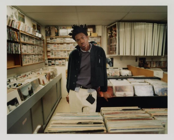 Young adult in a music store surrounded by rows of vinyl records and movies, wearing a dark jacket, light pants, and a beaded necklace.