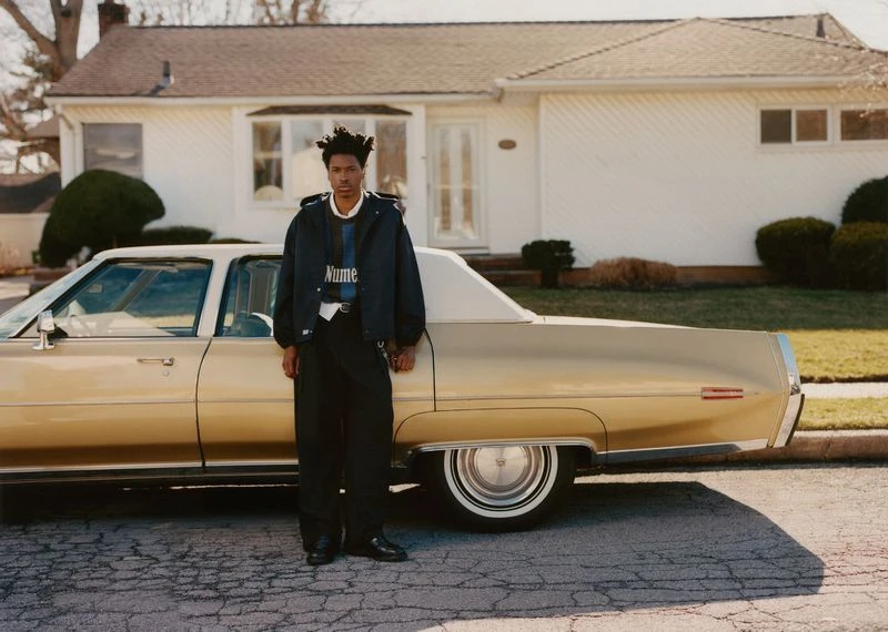 Young man in a navy jacket and black trousers stands next to a gold vintage car outside a white house with green lawn.