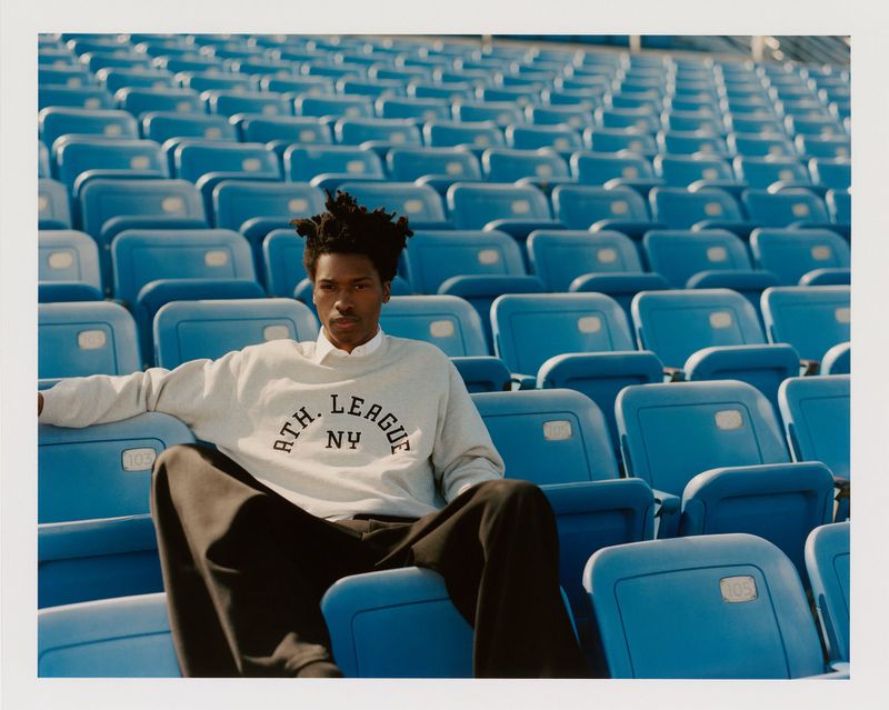A young Black man is seated in a stadium wearing a grey sweatshirt and black pants, showcasing fashion in an urban setting.
