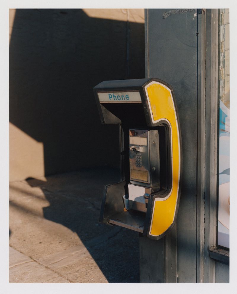 A yellow and black public payphone is mounted to the side of a building on a city street, with a shadow nearby.