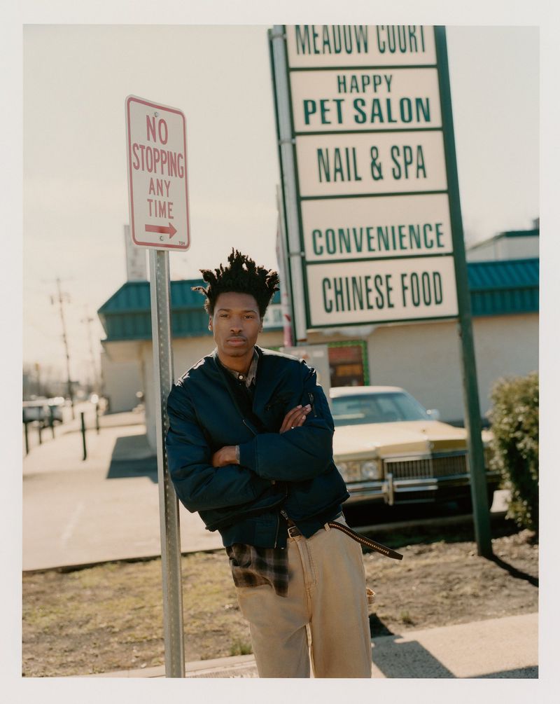 Man in blue bomber jacket and khaki pants poses near 'No Stopping' sign and a multi-business sign with a car in the background.