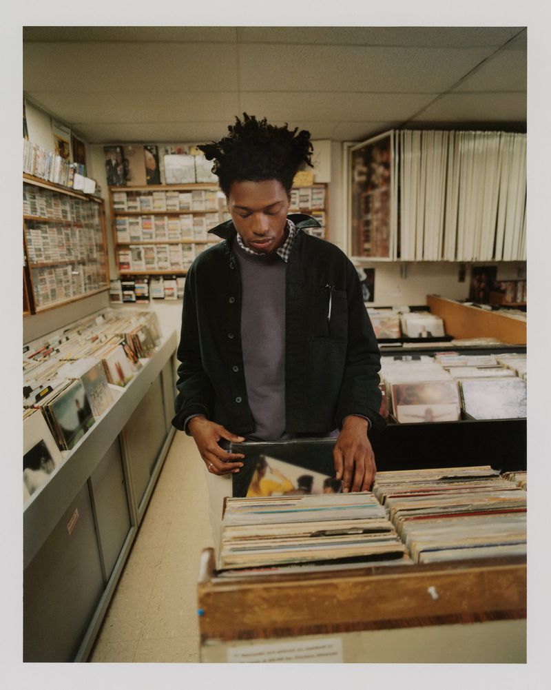 A young adult stands in a record store, browsing through vinyl records on a shelf in a softly lit interior setting.