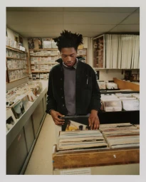 Young Black man with dreadlocks browsing vinyl records in a record store wearing a dark overshirt.