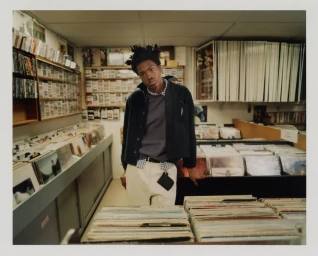 A young Black man wearing a dark jacket and cream trousers standing surrounded by vinyl records in a record store.