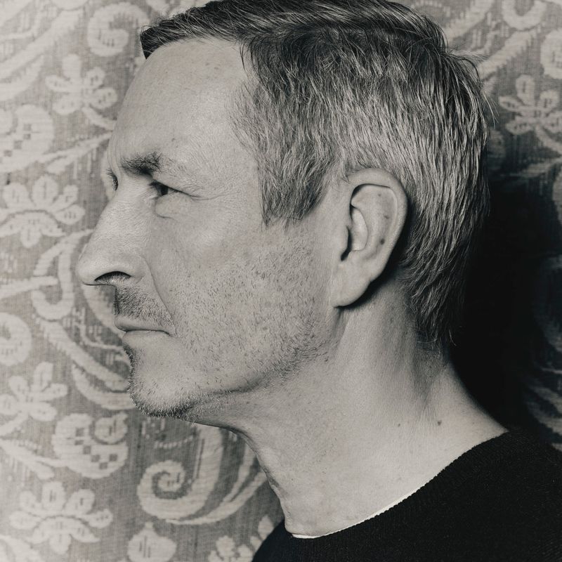 Close-up profile portrait of designer Dries Van Noten wearing a dark sweater against a woven damask textile backdrop.