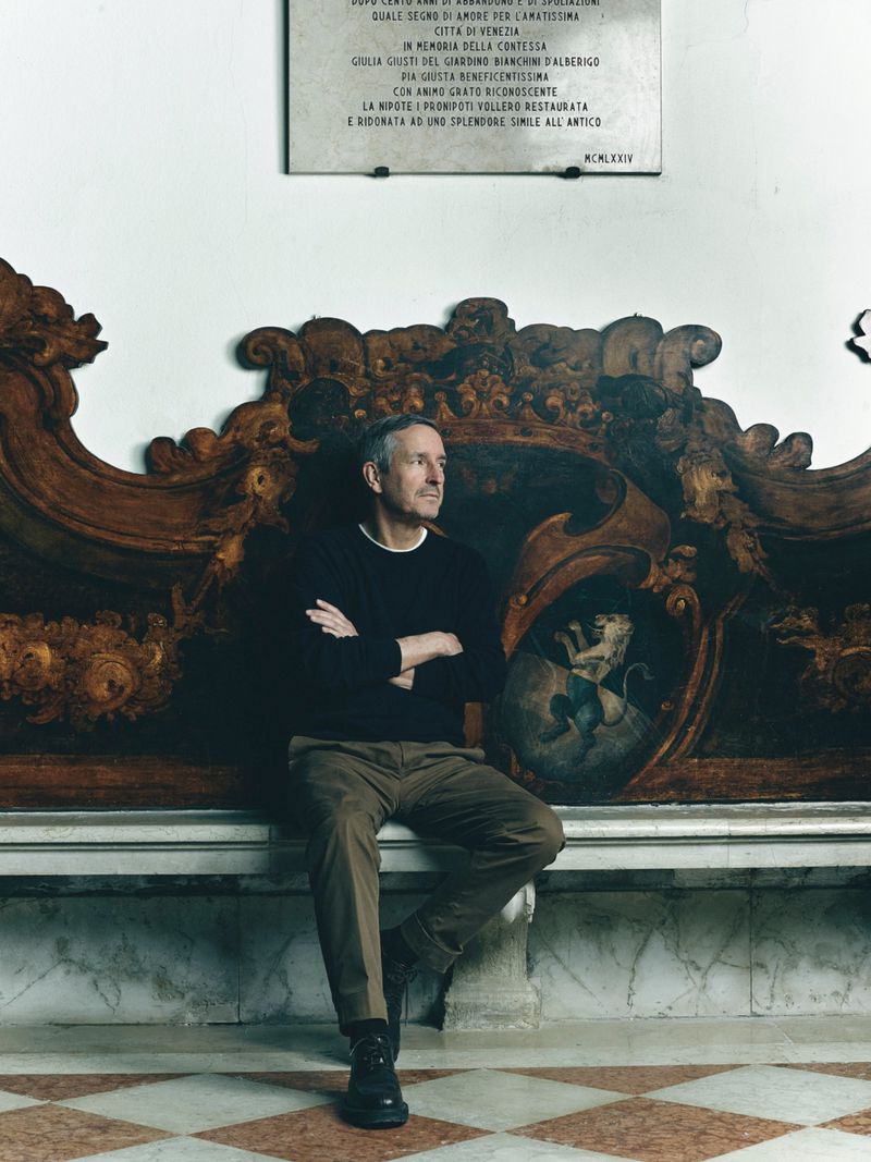 Man seated with arms crossed on an ornate wooden bench below a marble plaque on a pale wall