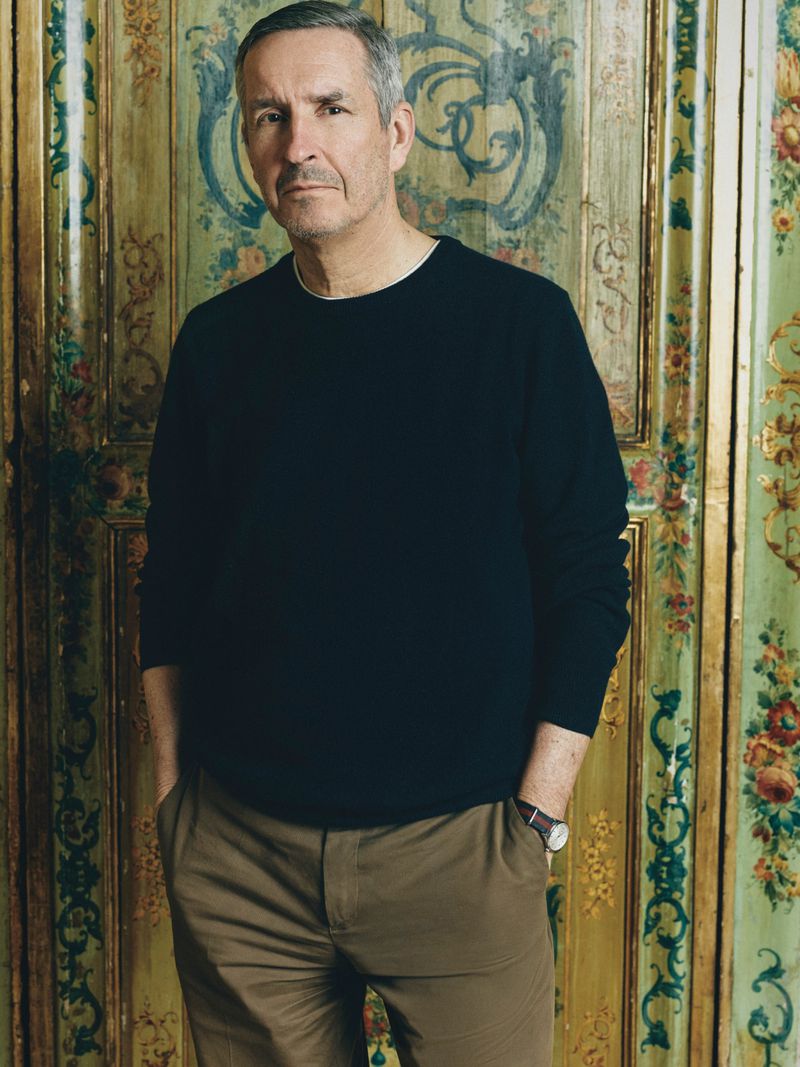 Portrait of fashion designer Dries Van Noten wearing a dark sweater and khaki trousers against an ornate painted screen.