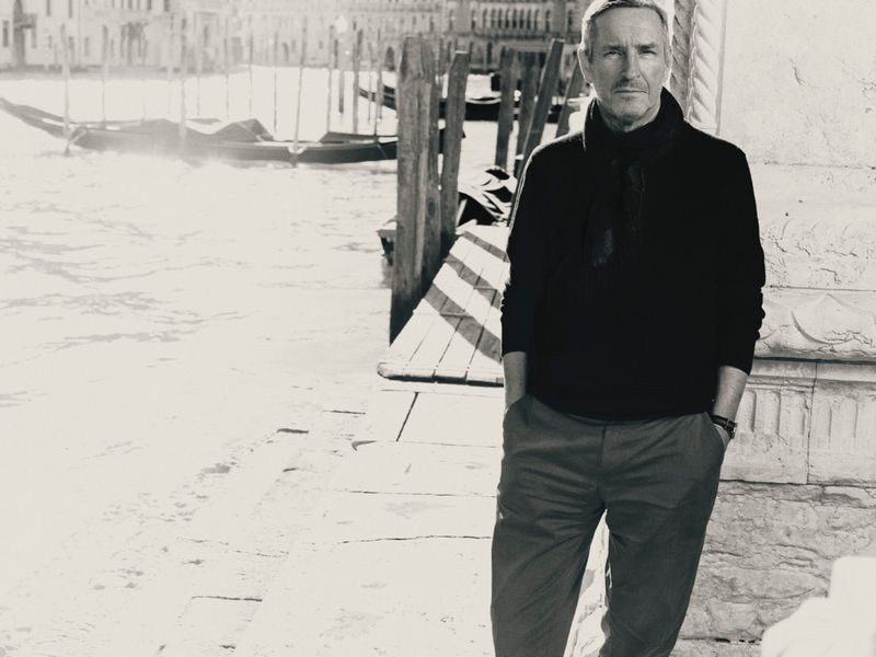 Black and white portrait of fashion designer Dries Van Noten standing beside a Venice canal with gondolas in the background.