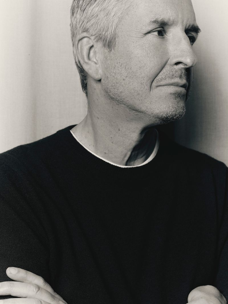 Black and white portrait of fashion designer Dries Van Noten in a black sweater with arms crossed against a neutral studio backdrop.