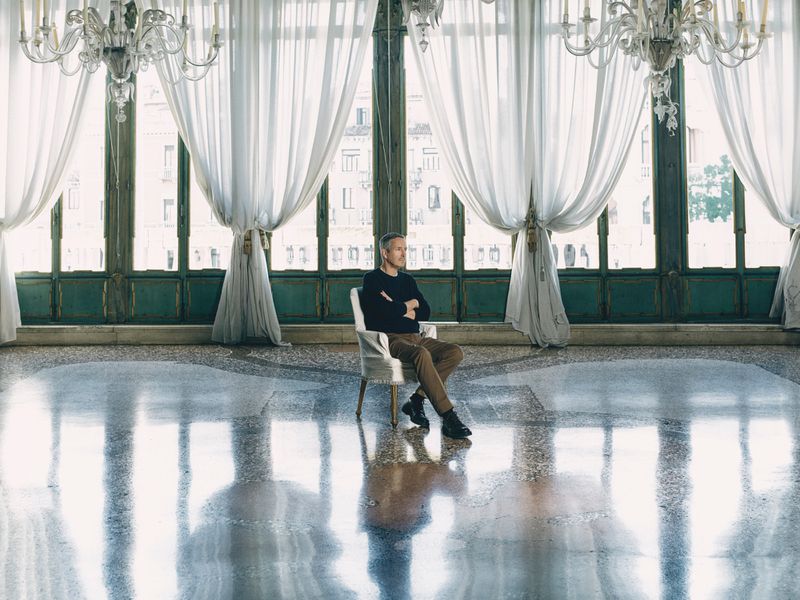 A man seated in a white armchair inside a grand hall with tall windows, sheer drapes, and ornate chandeliers.