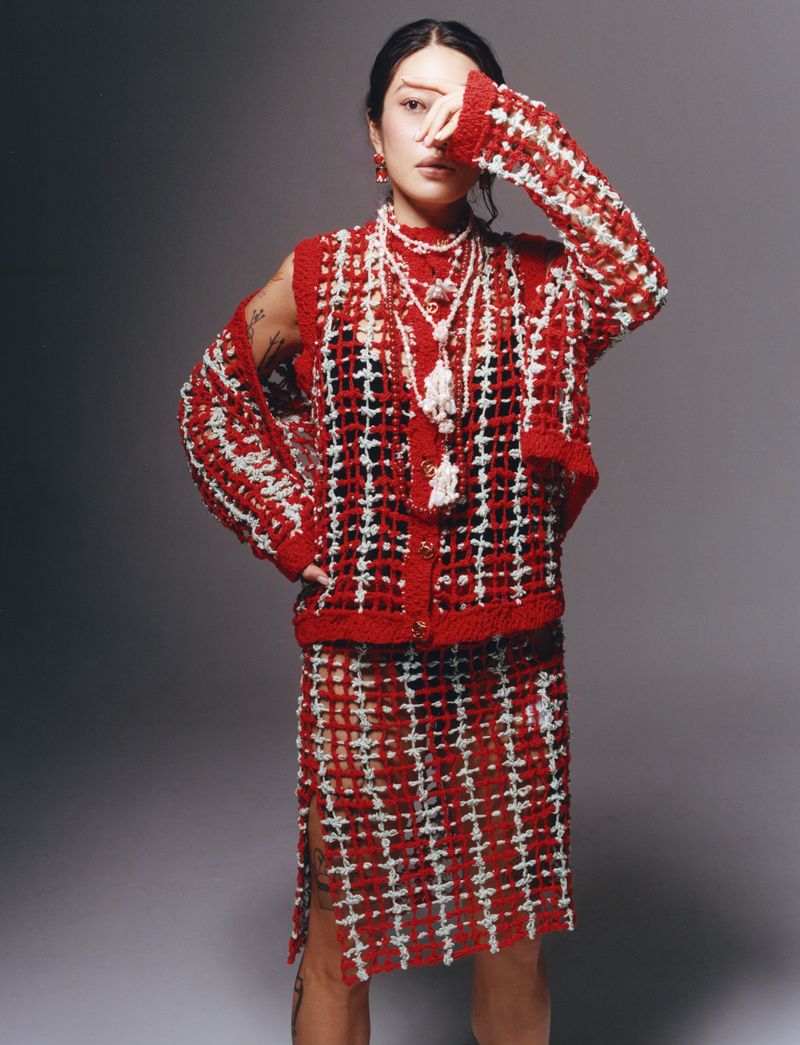 Model posing in a red and white open-knit patterned skirt set with beaded tassel necklaces against a grey studio backdrop.