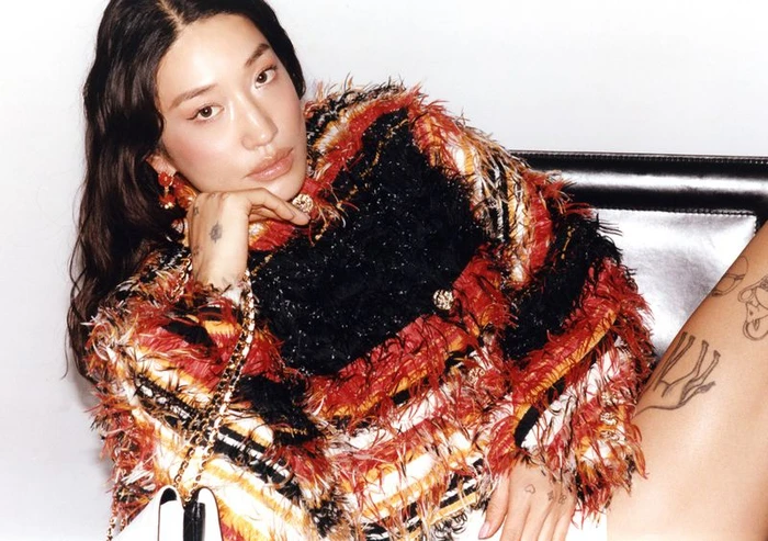 Peggy Gou poses in a textured red, black, and white fringed jacket while leaning against a black leather chair.