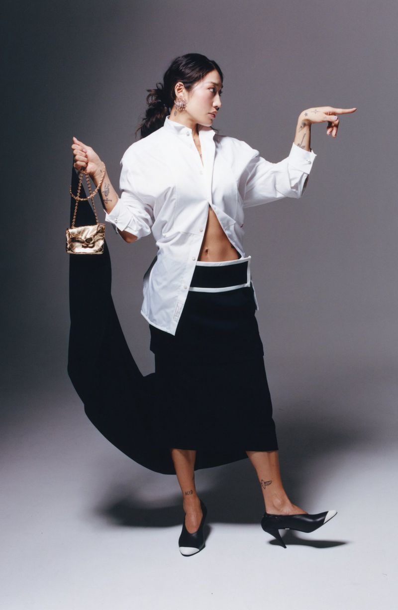 Peggy Gou poses in an open white shirt, black midi skirt, and two-tone heels holding a gold mini bag against a grey studio backdrop.