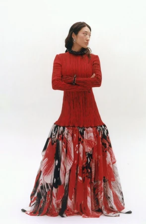 Woman posing with crossed arms wearing a long red dress with a painterly brushstroke skirt against a white studio backdrop.
