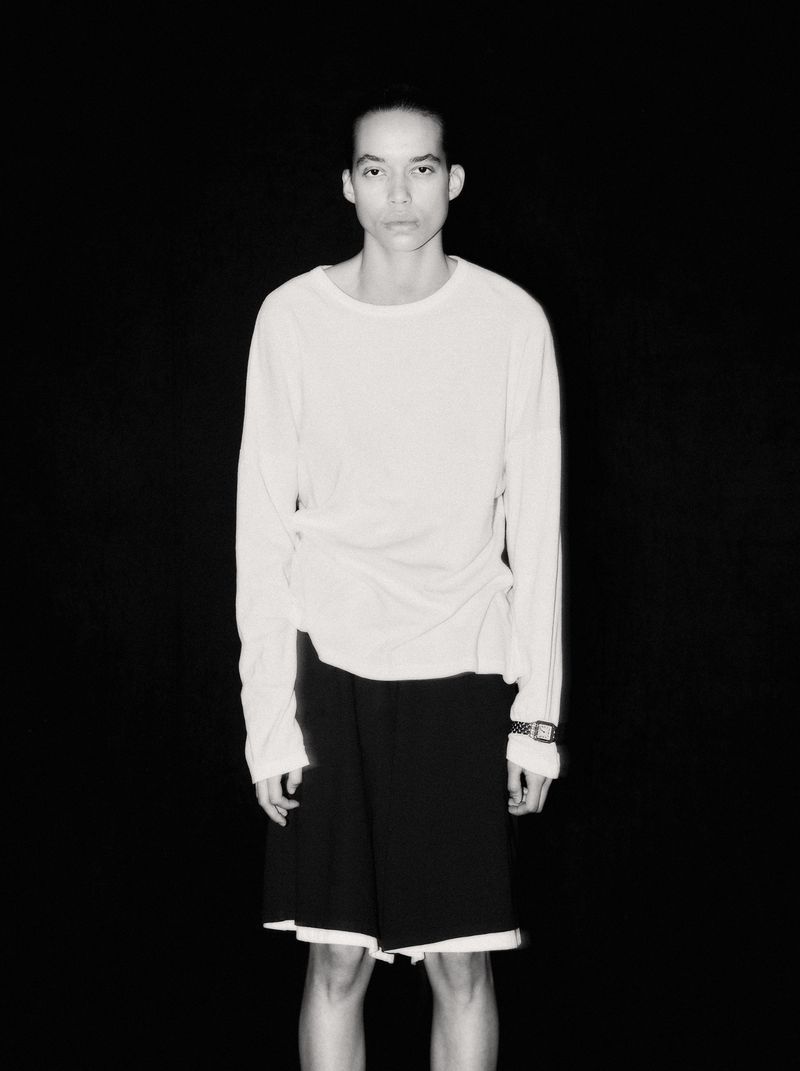 Lara Menezes wearing a white long-sleeve shirt and black shorts against a solid black background.