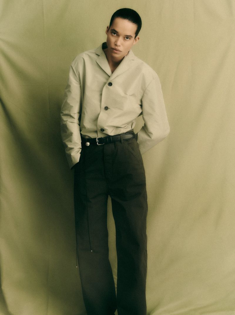 Model with short dark hair posing in a beige collared shirt and dark trousers against a draped olive fabric backdrop.
