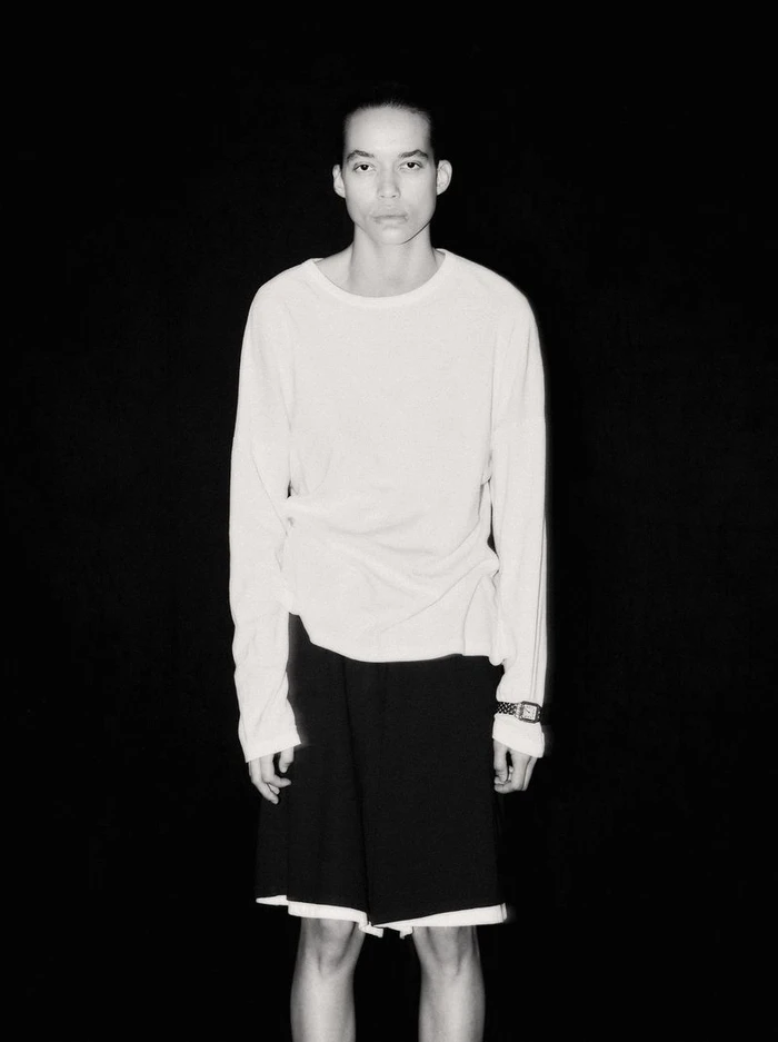 Lara Menezes wearing a white long-sleeve shirt and black shorts against a solid black background.