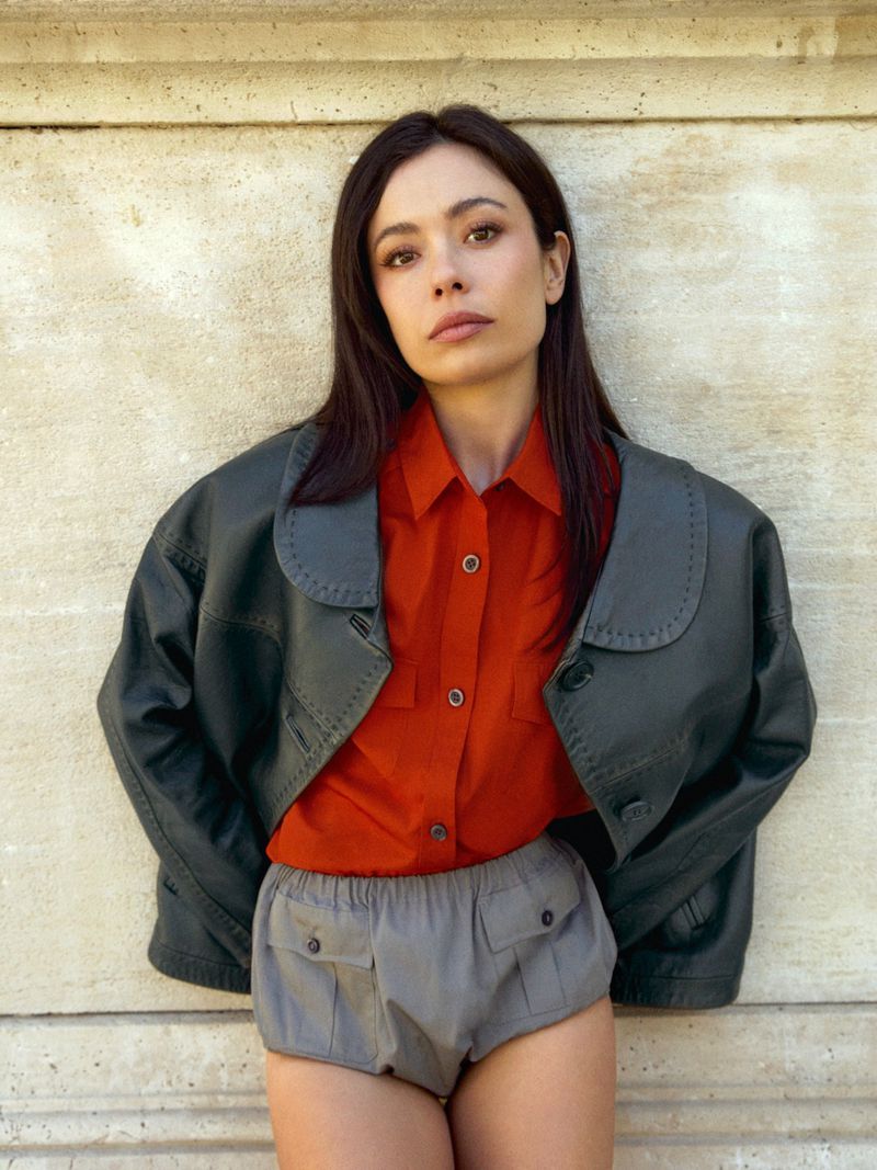 Actress Anna Castillo wearing an orange shirt, grey pleated shorts, and an oversized dark leather jacket against a stone wall.