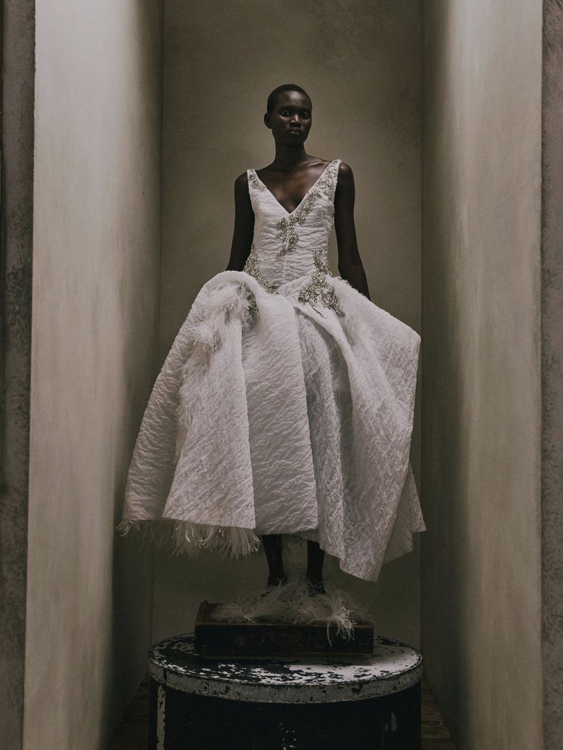 Model Aluel Keror posing in an embellished white textured gown while standing on a pedestal inside a neutral architectural alcove.