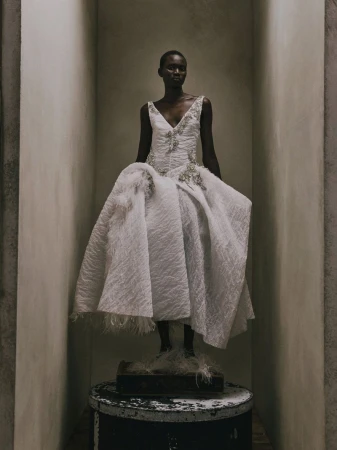 Model Aluel Keror posing in an embellished white textured gown while standing on a pedestal inside a neutral architectural alcove.