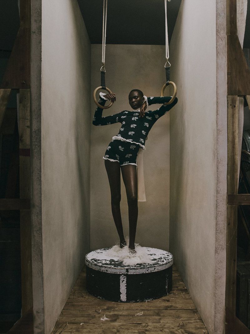 Model Aluel Keror standing on a round pedestal holding gymnastic rings in a dark patterned knit top and shorts for Erdem.