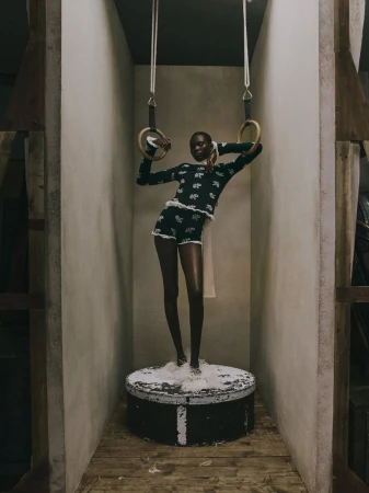 Model Aluel Keror standing on a round pedestal holding gymnastic rings in a dark patterned knit top and shorts for Erdem.
