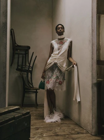 Model in embroidered floral dress standing against a neutral studio wall with stacked wooden chairs