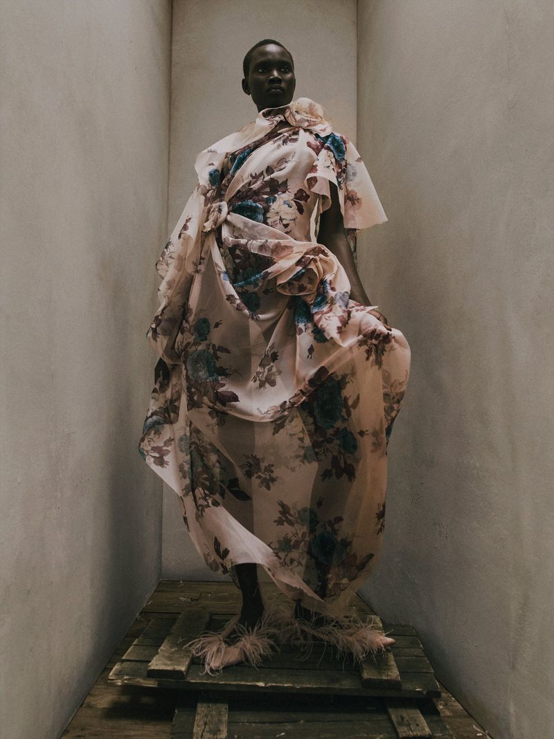 Model Aluel Keror stands in a sheer draped floral gown and feathered shoes inside a minimalist concrete set.